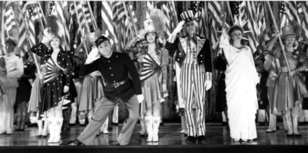 A line of performers on a stage in patriotic, flag-themed costumes (including Uncle Sam) saluting with hands to their foreheads, American flags in the background.