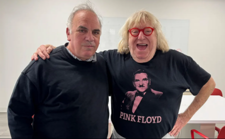 2026-07-08_13-13-41 - We Got Bruce! Two men pose together with arms around each other in a bright room; left man in a dark sweater, right man wearing a Pink Floyd shirt and red glasses, smiling.
