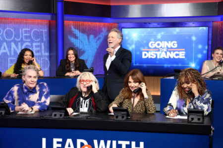 Project Angel Foods Lead With Love 7 - A Fundraising Special On KTLA - We Got Bruce! A television panel show set with five people at a long desk, a host standing and gesturing, and a screen behind reading 'GOING THE DISTANCE'. The panelists use phones to speak into headsets as cameras capture the moment.