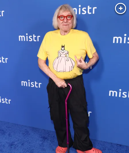 2026-10-29_04-17-40 - We Got Bruce! Person with blonde bob and red glasses wearing a yellow shirt with a princess graphic, black pants, pink cane, and orange clogs on a blue backdrop.